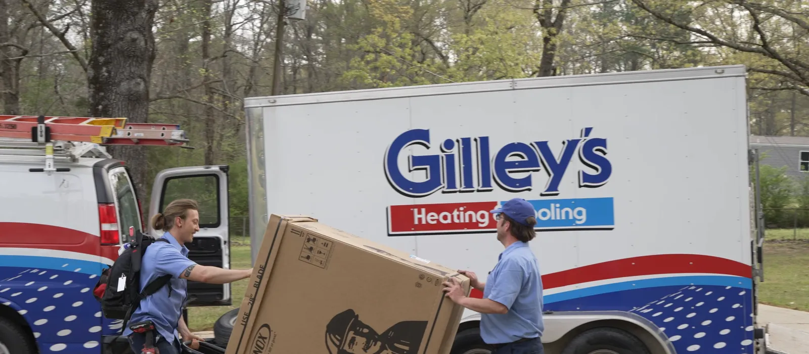 Two Gilley's installers wheel a new outdoor unit on a dolly past the Gilley's trailer
