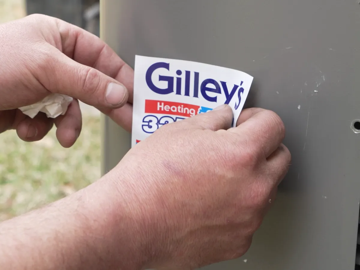 A hand presses a Gilley's Heating and Cooling sticker onto a new outdoor unit