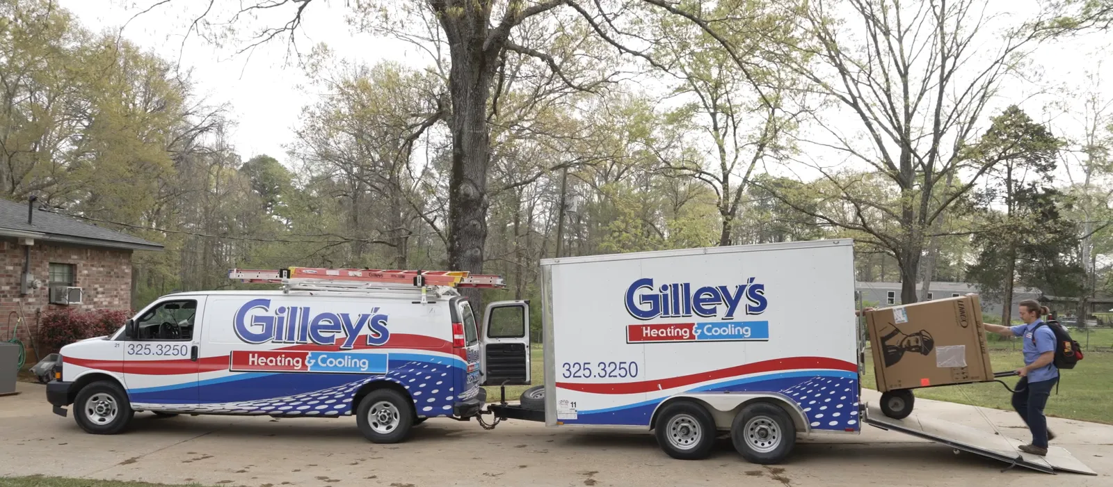 A Gilley's van and trailer at a customer's home while an installer wheels a new unit up the drive